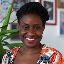 A person with braided hair wearing a patterned orange and white top stands indoors in front of a green plant and framed pictures on the wall.