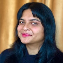A person with long dark hair wearing a dark top is photographed in front of a softly lit golden curtain background. They are faintly smiling and looking to camera.
