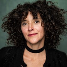 Head and shoulder photo of Anna Seymour, a person with curly brown hair wearing a dark top photographed in front of a softly lit green background. They are faintly smiling and looking to camera.