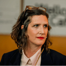 Head and shoulder photo of Laurie Britton Newell, a person with brown hair wearing a black blazer and white collared shirt. They are looking to their left.