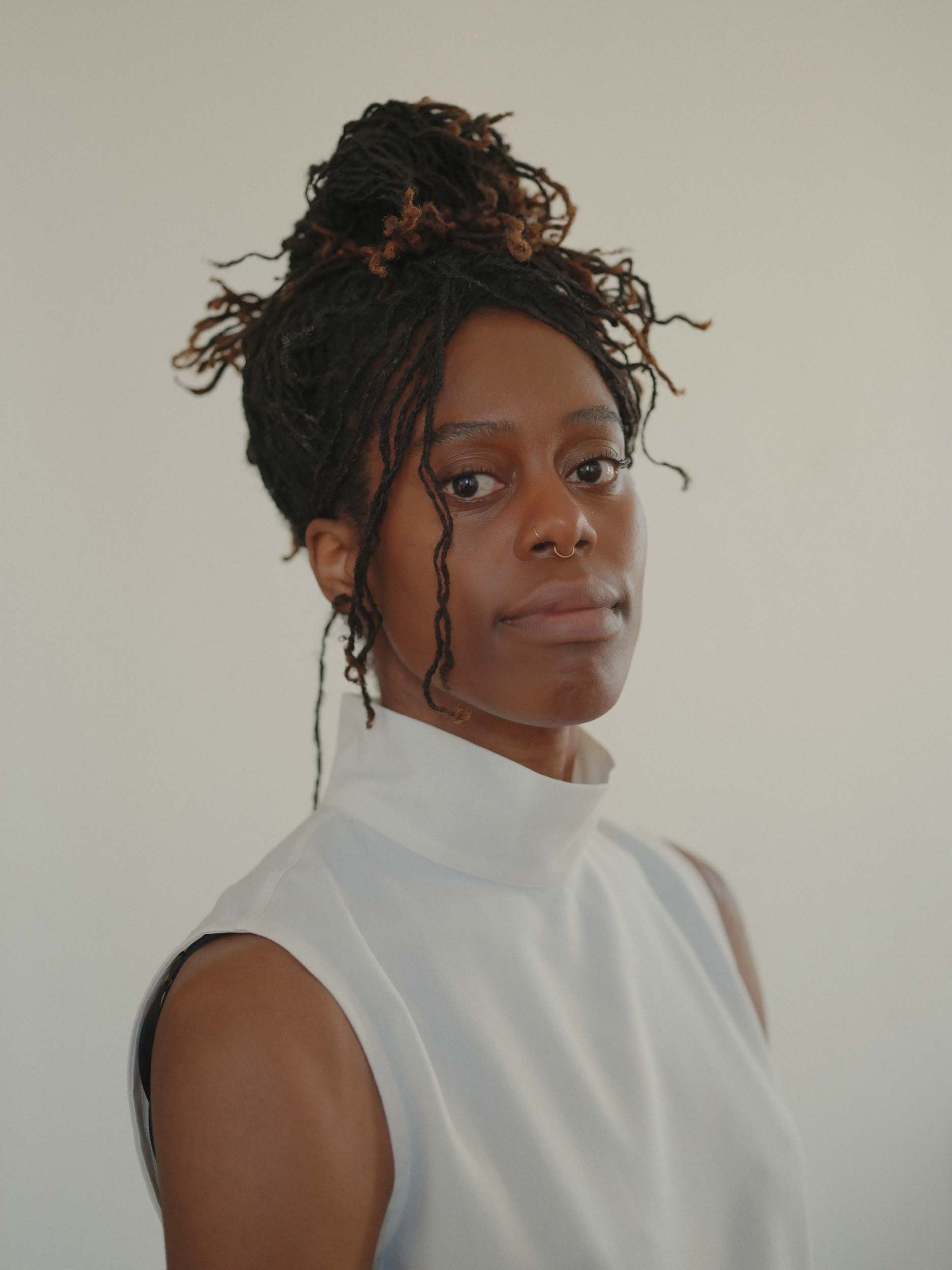 Photo of Temitope Ajose, a person with dark hair in a high bun, wearing a white high-neck vest. They are looking to camera, with their body angled to the side.
