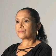 Portrait photograph of a woman in a black top with criss-cross straps and gold hoop earrings, hair pulled back, standing against a plain light grey background.
