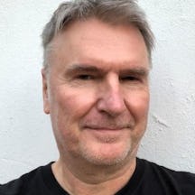 A headshot photo of a man with grey hair wearing a black top