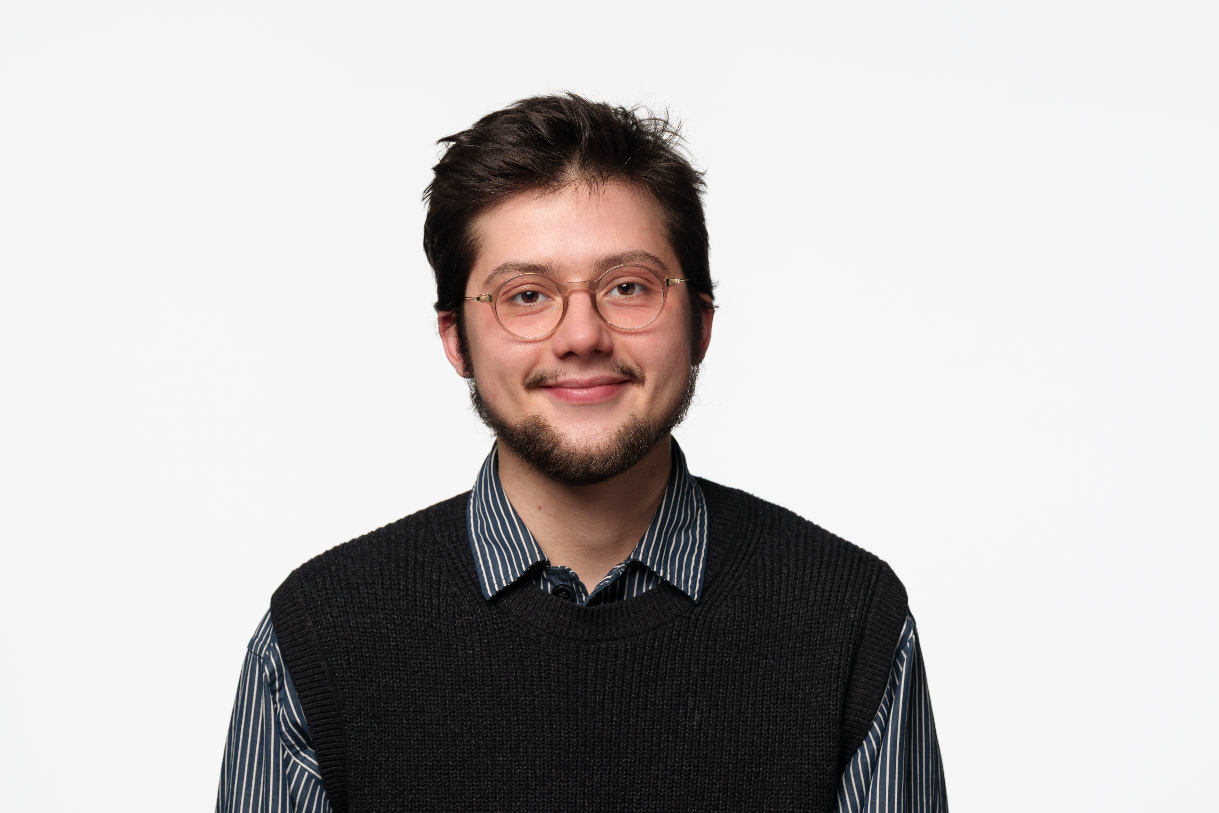 A person with short dark hair is facing the camera against a plain white background, wearing a dark knitted vest over a striped button-up shirt.