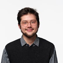 A person with short dark hair is facing the camera against a plain white background, wearing a dark knitted vest over a striped button-up shirt.