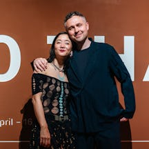 A photo of Christine Sun Kim (left) and Thomas Mader (right) embracing, looking at the camera. Taken in front of a large sign with details of their 1880 THAT exhibited on it.