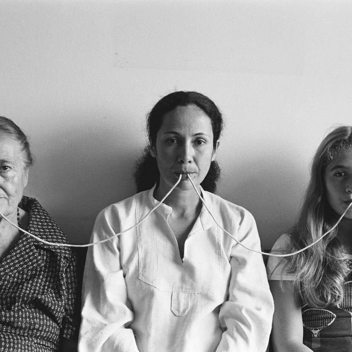 Anna Maria Maiolino wears a white shirt and sits between her elderly mother and her daughter. They are connected at the mouth by a thread.