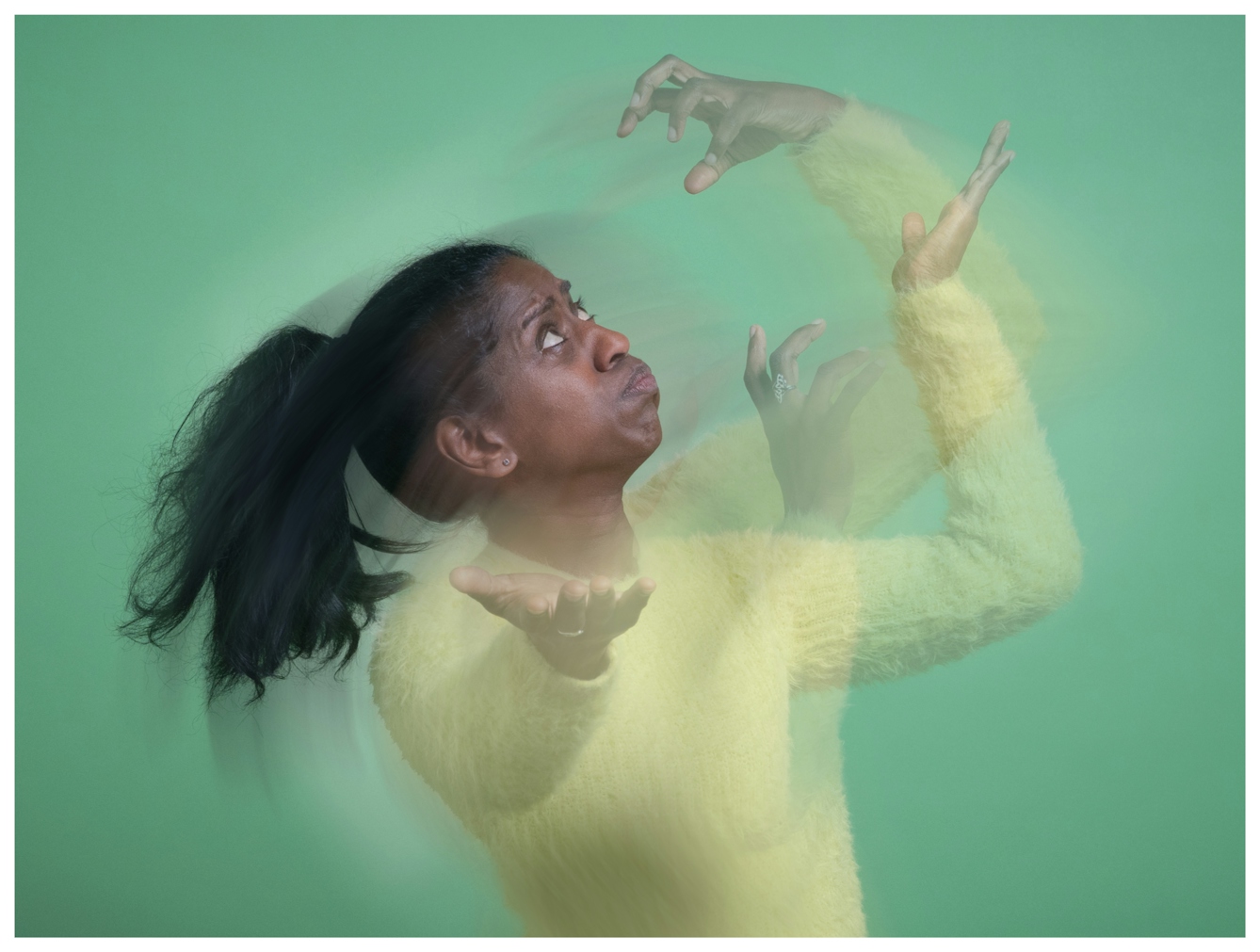 Portrait of Nadia Nadarajah in a yellow jumper posing against a green background. Her arms create a blurred motion effect, expressing dynamic energy and intensity.