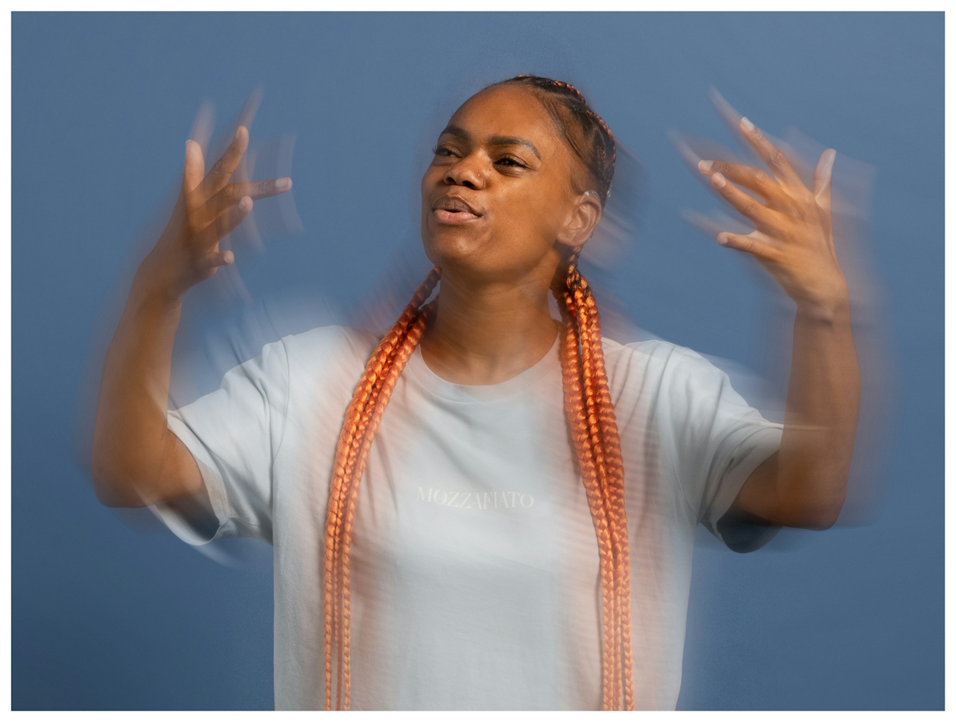 Portrait of Sarah Adedeji with long, orange braids animatedly gesturing with blurred motion against a blue background, as she signs the lyrics to a song.