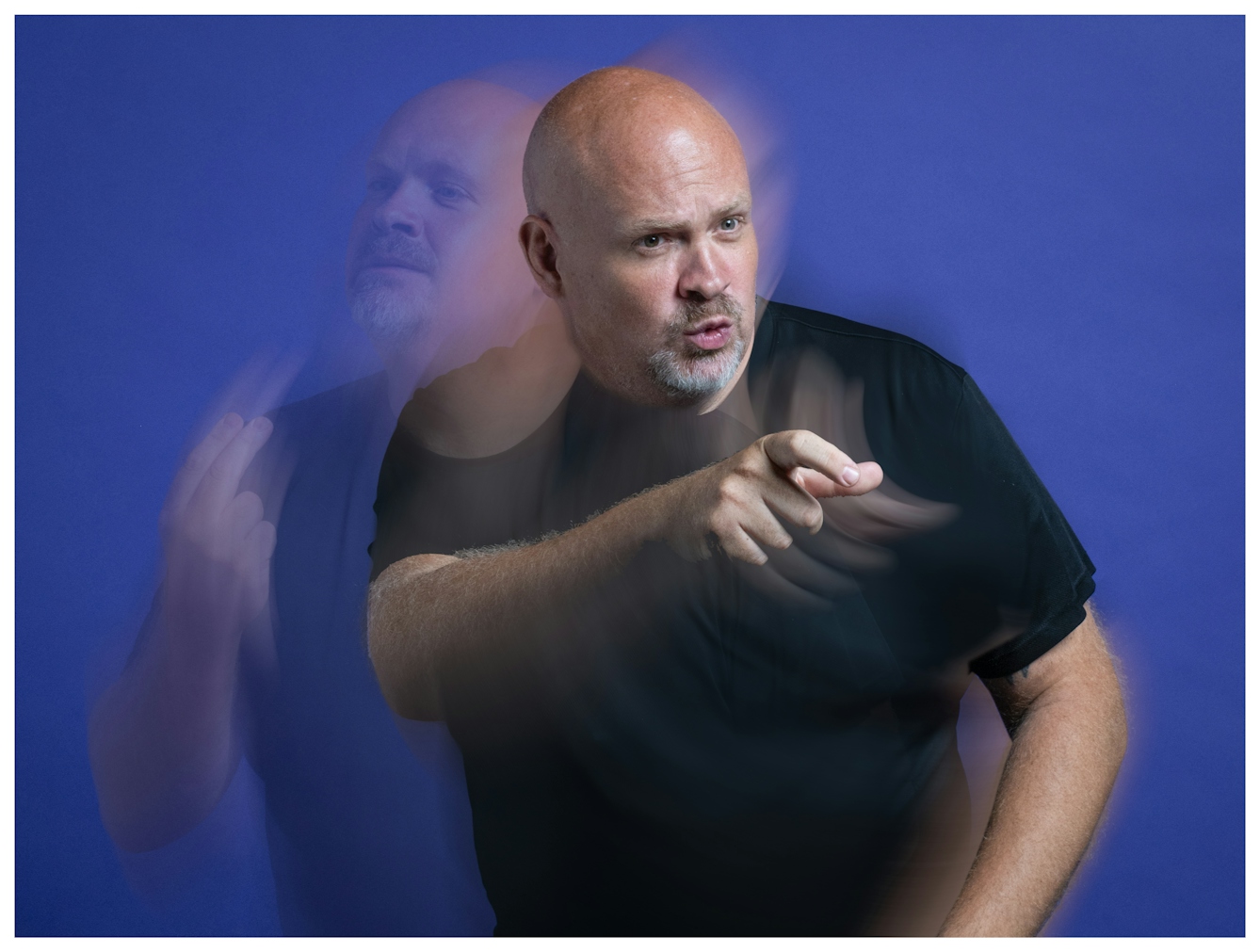 A portrait of comedian Gavin Lilley, a bald man in a black shirt pointing forward, with motion blur effects showing multiple positions of his arms and head against a blue background.