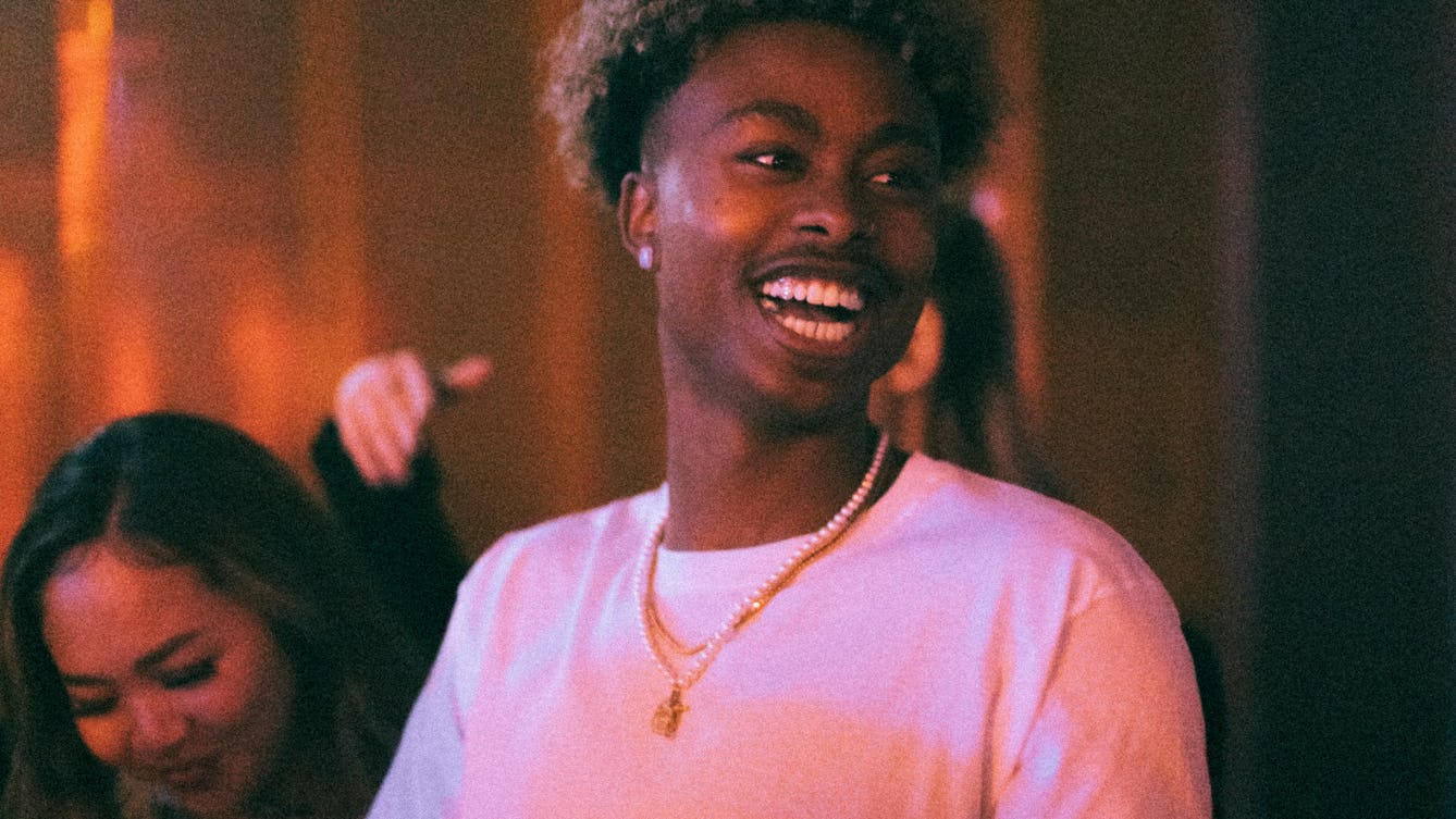 A young person looks off camera with a large smile on his face. There are people in the background, and the lights are giving a pink hue.