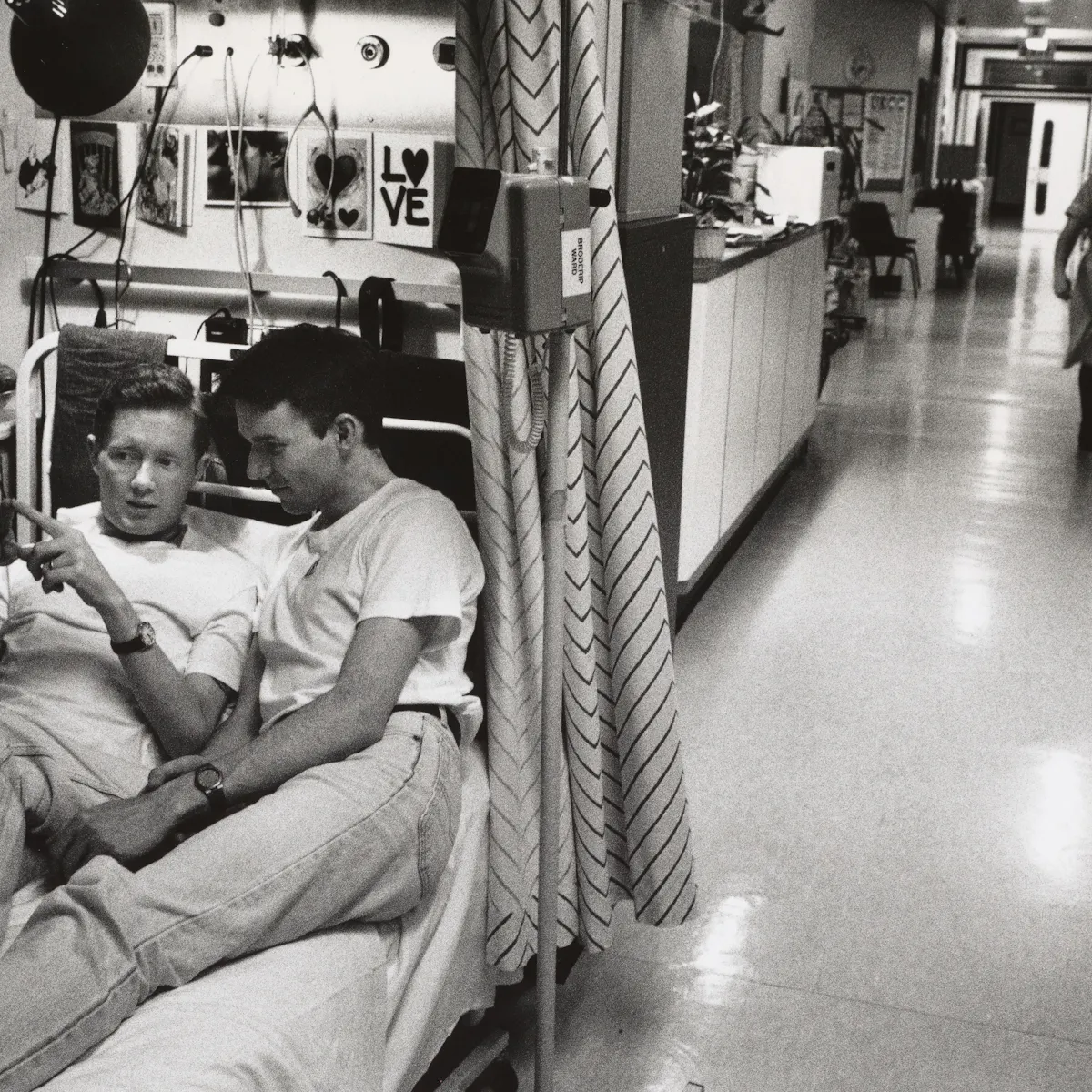 Black and white photo of a hospital ward, with two people in casual clothes lying together on a narrow hospital bed in the foreground. The bed is surrounded by medical equipment, drinks and personal items, including photos and cards on the wall. In the background, a nurse walks alone down a long corridor lined with curtains.