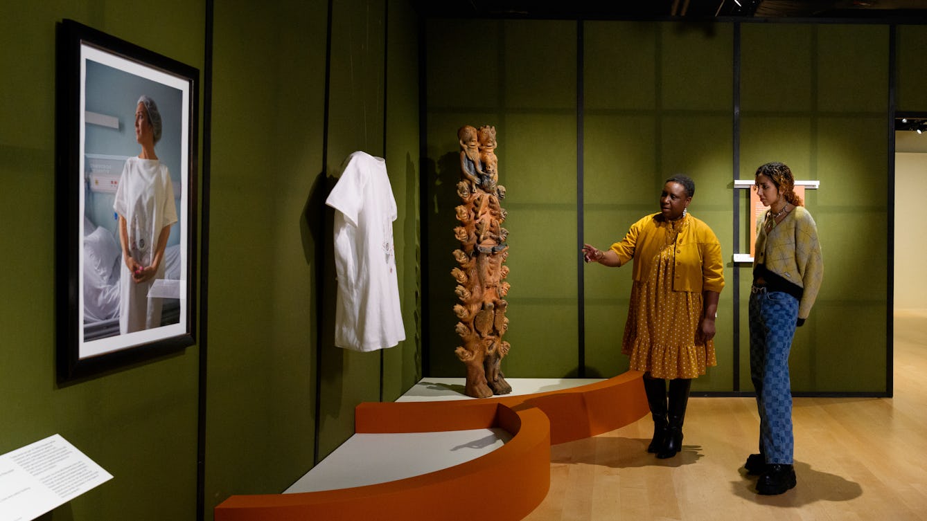 Gallery visitors looking at a tall clay sculpture, embroidery textiles and photography.