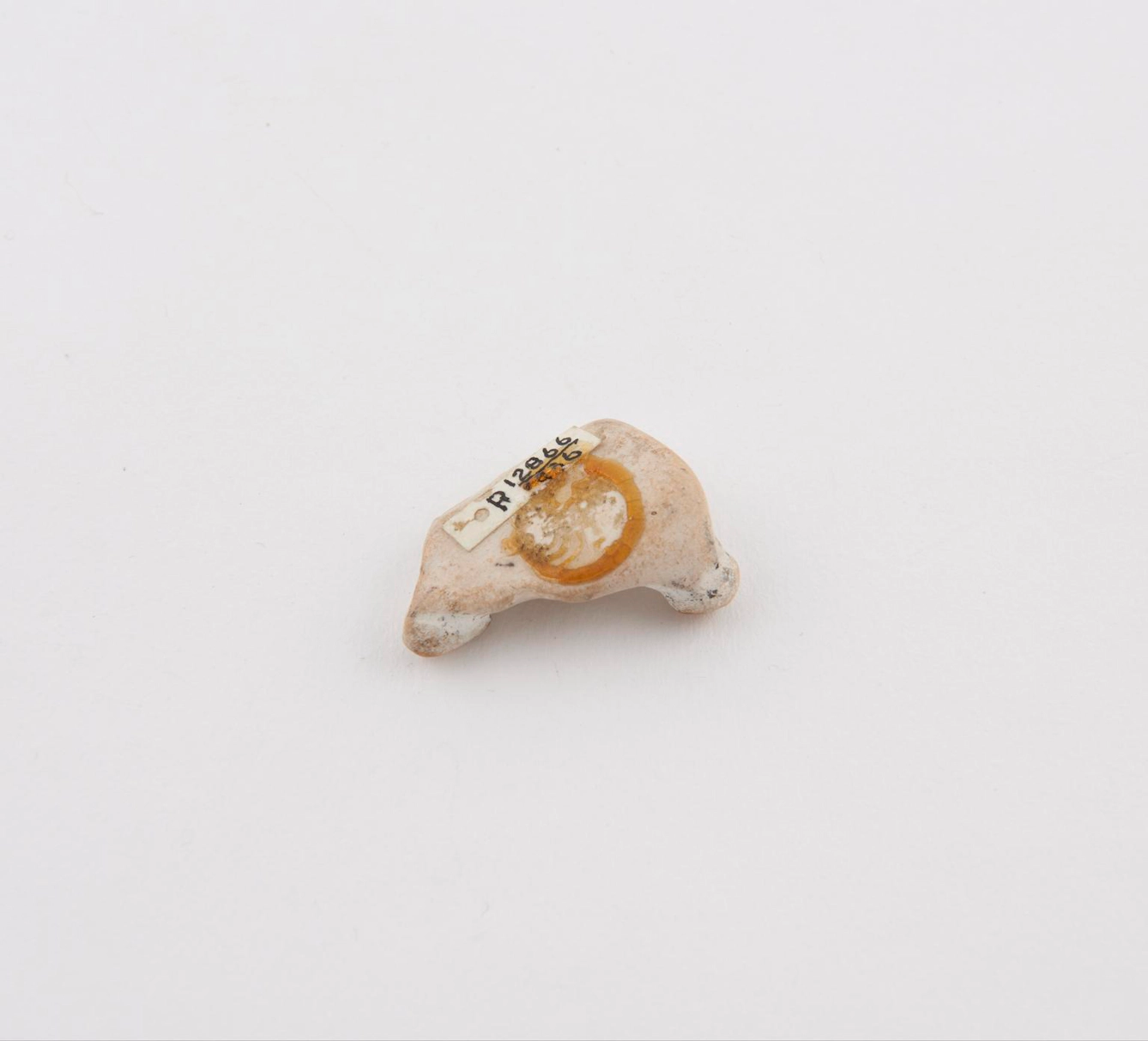 A small, worn, pale piece of flint is shown on a plain white background, with a circular yellow glaze on its side and a tiny catalog label with handwritten numbers attached to the top.