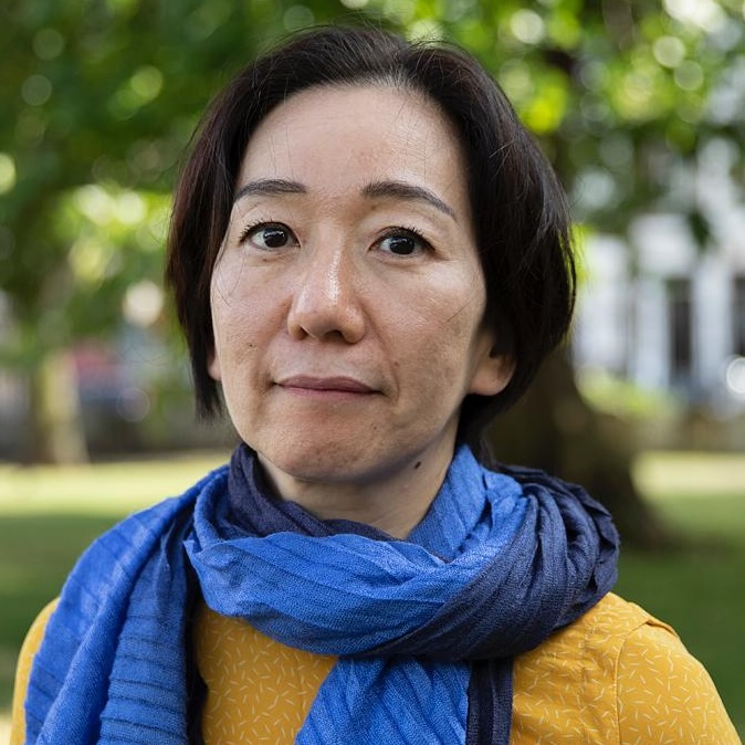 A person with short dark hair is standing outdoors in a sunlit park, wearing a mustard-yellow top and a blue scarf wrapped around their neck.