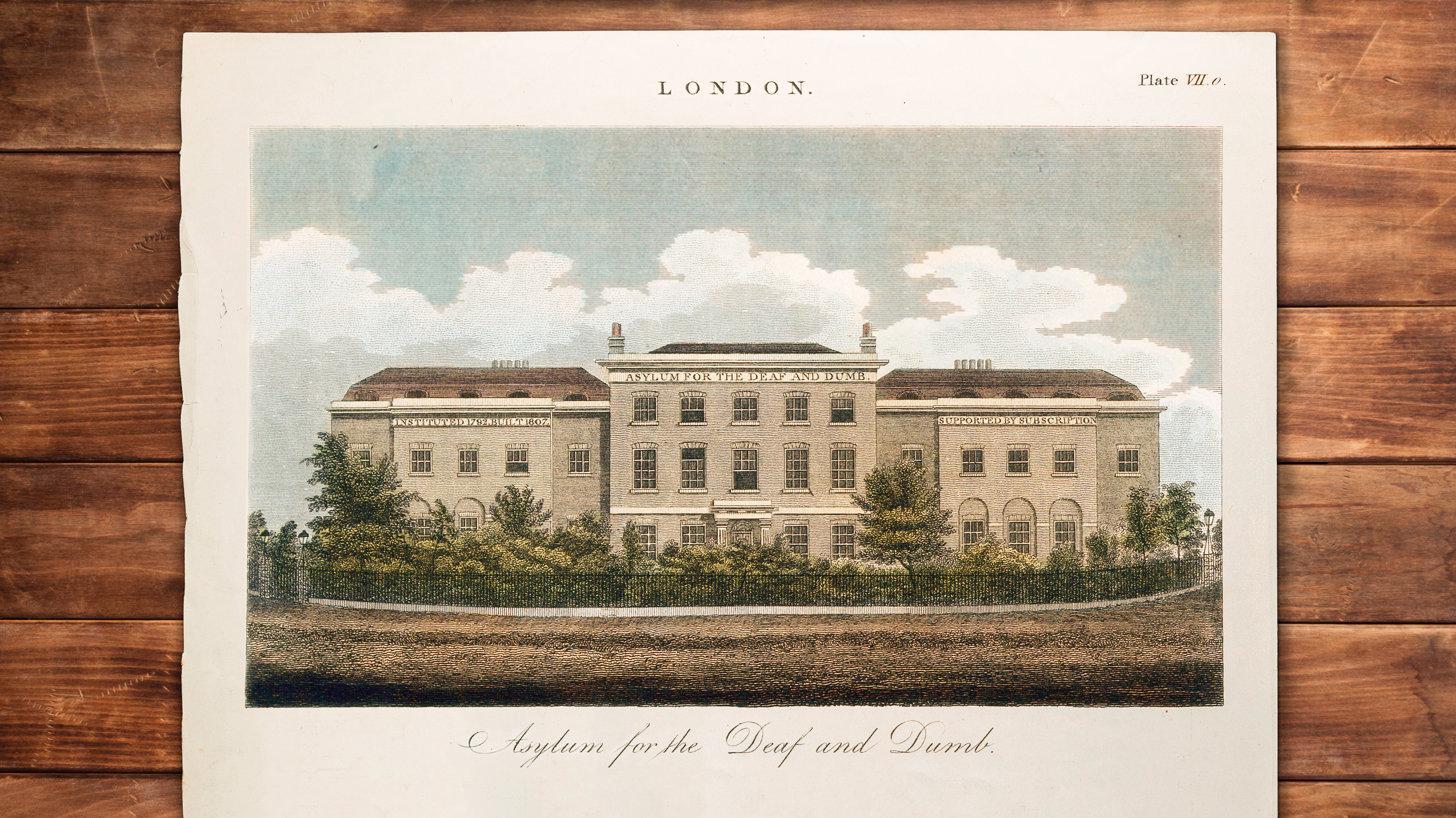 Wooden tabletop on which rests a colour engraving of a large 19th century stone building. Lettering on the front of the building reads "Asylum for the Deaf and Dumb."