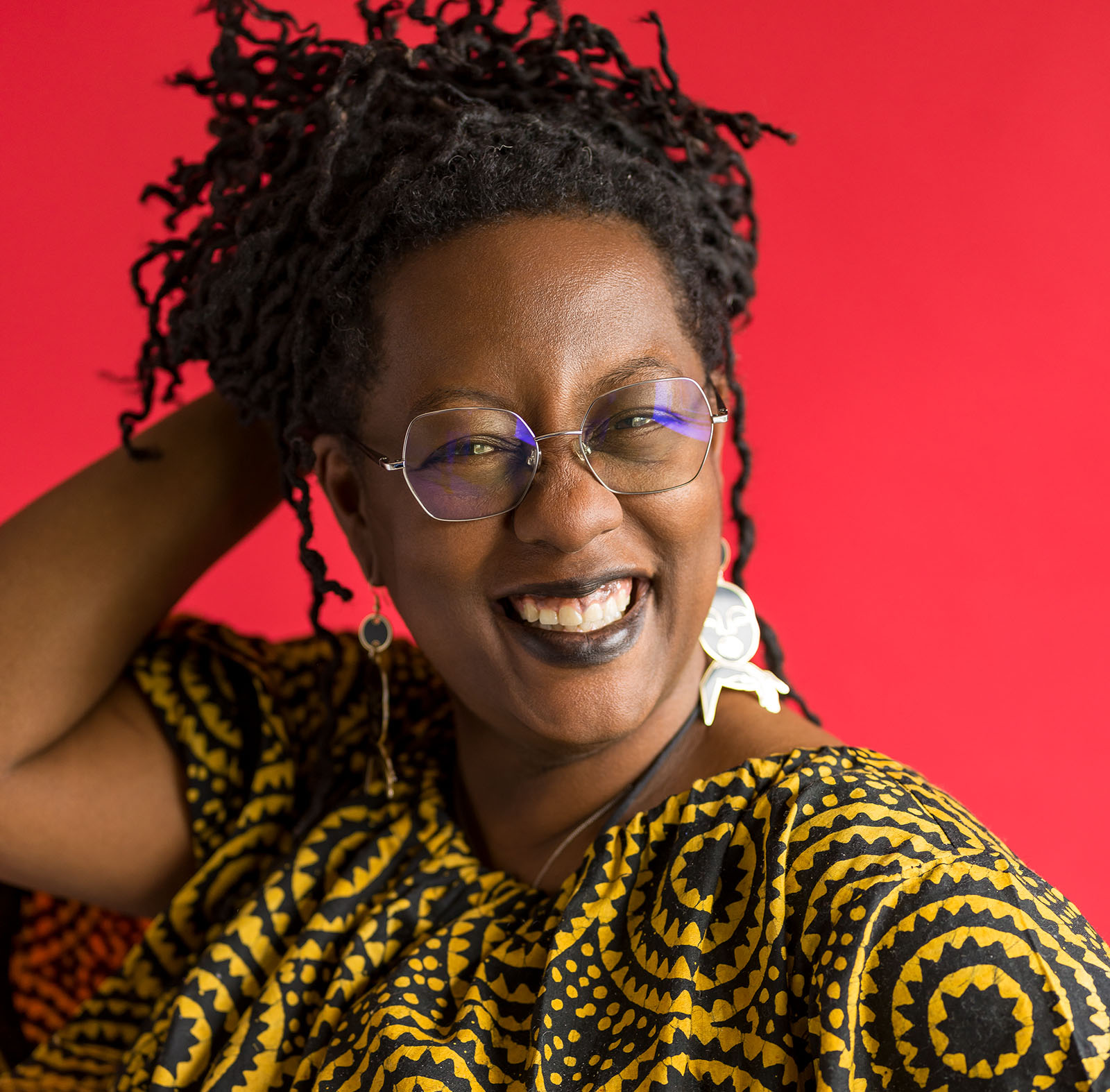 Black and white photo of the head and shoulders of a woman wearing glasses and dangling earrings. She has a broad smile and is looking directly at the viewer.