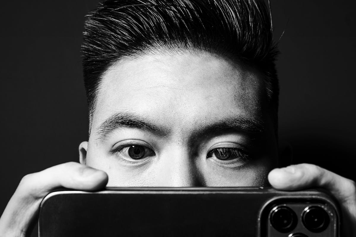 Black and white photographic portrait of a close-up of a man's face. He is holding up a smartphone close to his eyes such that it obscures his nose and mouth. His eyes are directed towards the phone's screen. His finders are visible holding the phone on either side and the camera lenses of the phone appear in the bottom right corner of the image. The man's face and hands are spotlit in a small circle of light. He is standing against a black background which means he is surrounded by darkness.