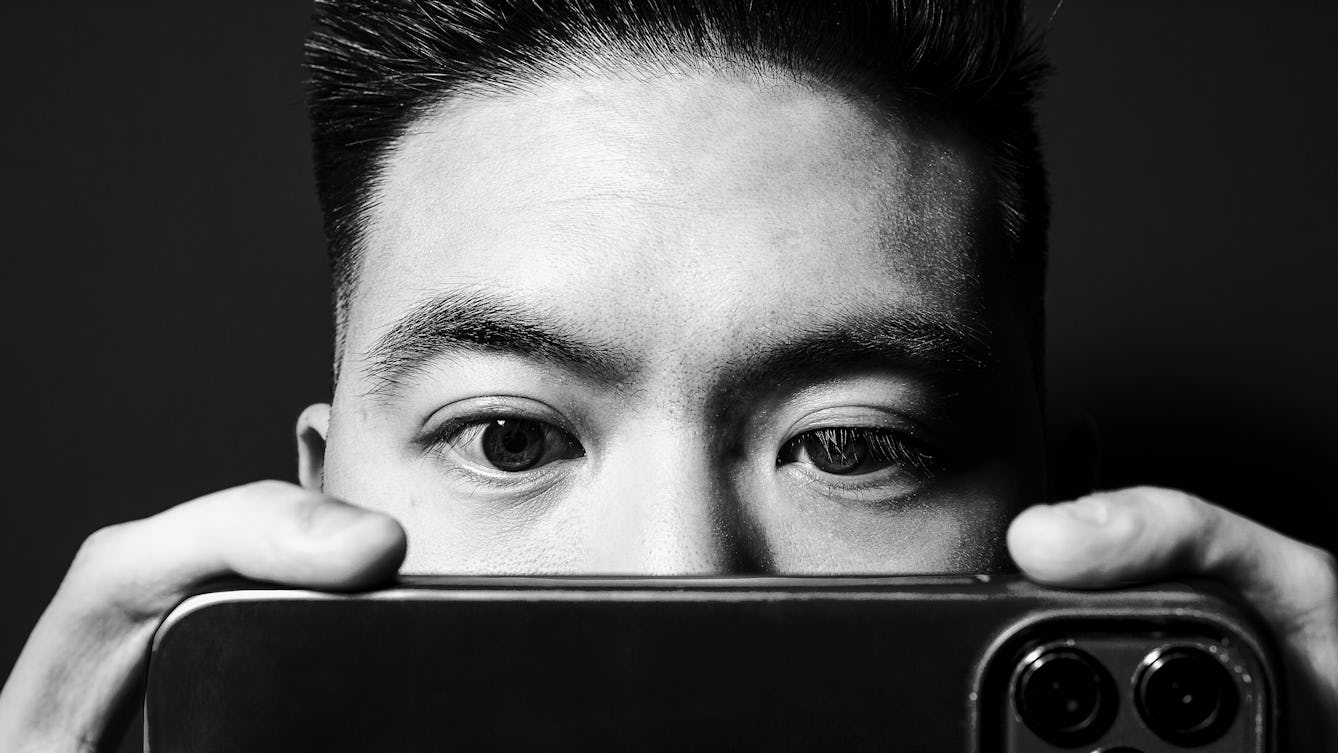 Black and white photographic portrait of a close-up of a man's face. He is holding up a smartphone close to his eyes such that it obscures his nose and mouth. His eyes are directed towards the phone's screen. His finders are visible holding the phone on either side and the camera lenses of the phone appear in the bottom right corner of the image. The man's face and hands are spotlit in a small circle of light. He is standing against a black background which means he is surrounded by darkness.