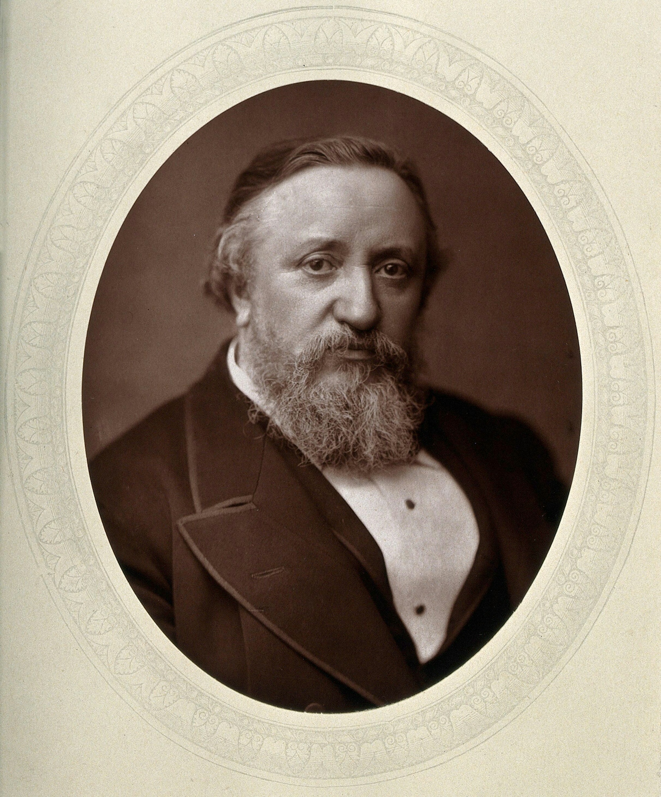 Black and white photographic portrait, showing the head, shoulders and chest of a man, who is looking straight ahead. He has short, greying hair and a beard. He is wearing a black suit jacket with a white shirt.