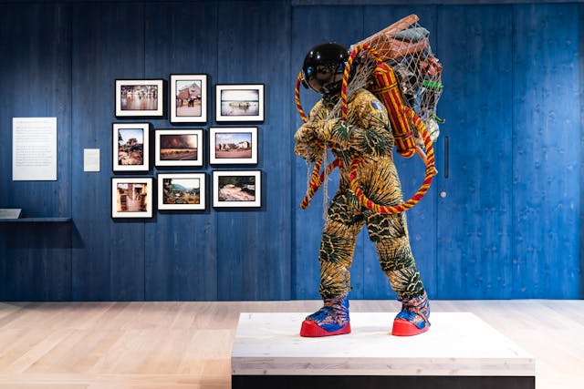 Photograph of an exhibition gallery space, with a blue stained wood wall in the background, on which are hung 9 photographs showing flooded landscapes. In the foreground is a life-size artwork of a figure resembling an astronaut. carrying a large net containing assorted objects including a suitcase.