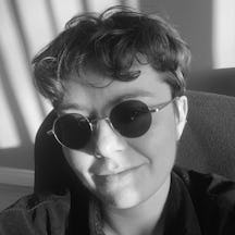 Black and white photograph of the head and shoulders of a young, white person. The person has short hair and is wearing round sunglasses. 
