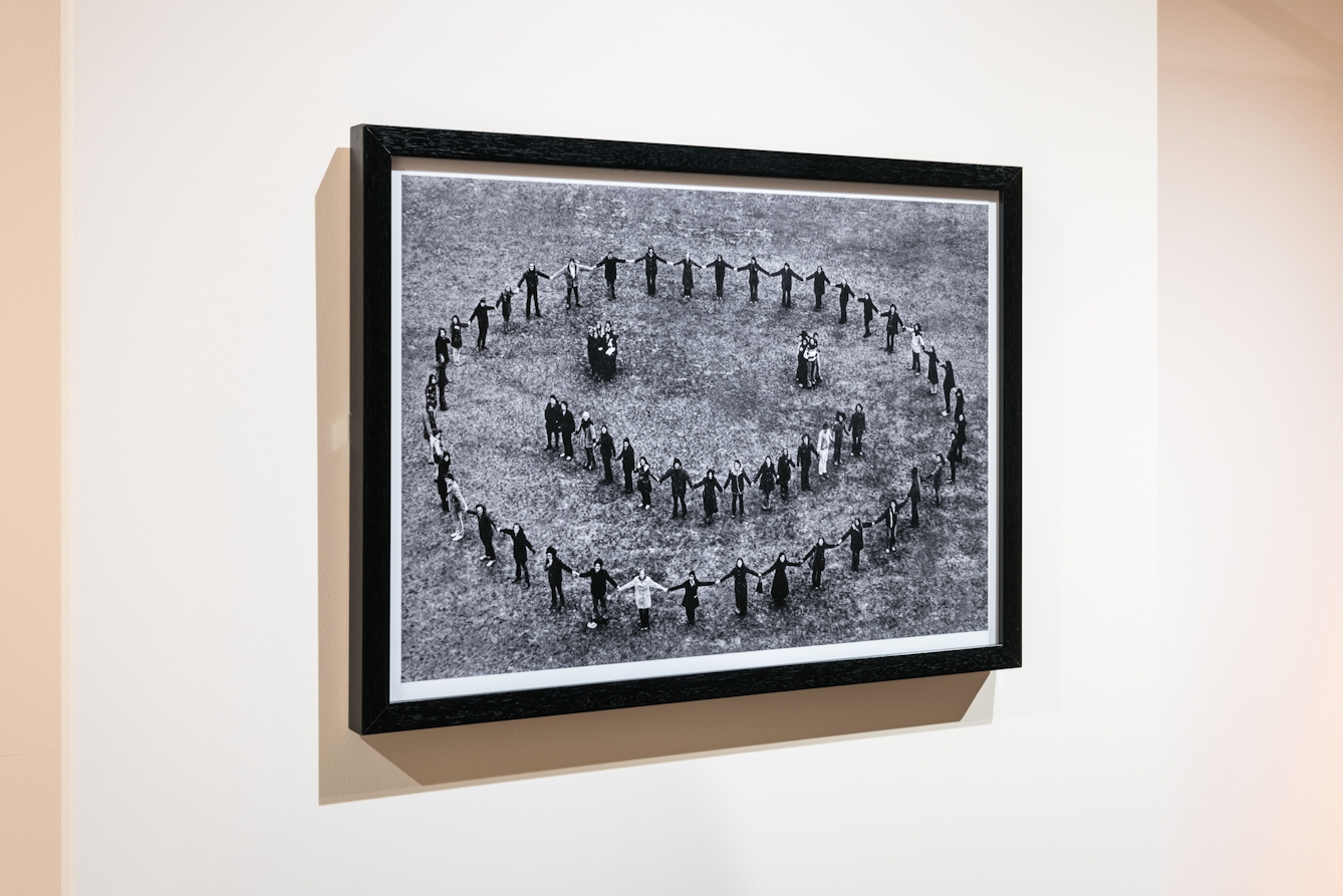 Photograph of an exhibition gallery showing a framed print hung on a cream coloured wall. The frame is black and mounted inside is a black and white photograph showing an aerial view of people on the ground holding hands and forming a circle, within which are clusters of people representing two eyes and a mouth. Together they all make a large smilie face.