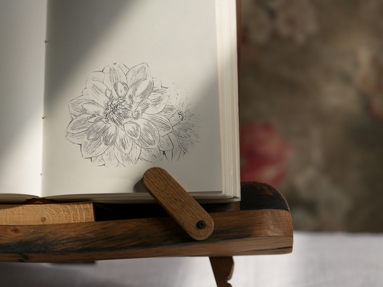 Photograph of a notebook on a wooden book stand, lit by a window from the right, casting a shadow on half of the page. On the right page there is a sketch of a flower.