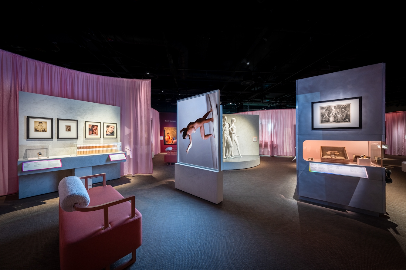 Photograph of a wide gallery view showing display cases, benches and wall mounted artworks. At the centre is a double sided screen showing the projected image of a morphing human body. In this image the projected human figure is rotated 90 degrees.