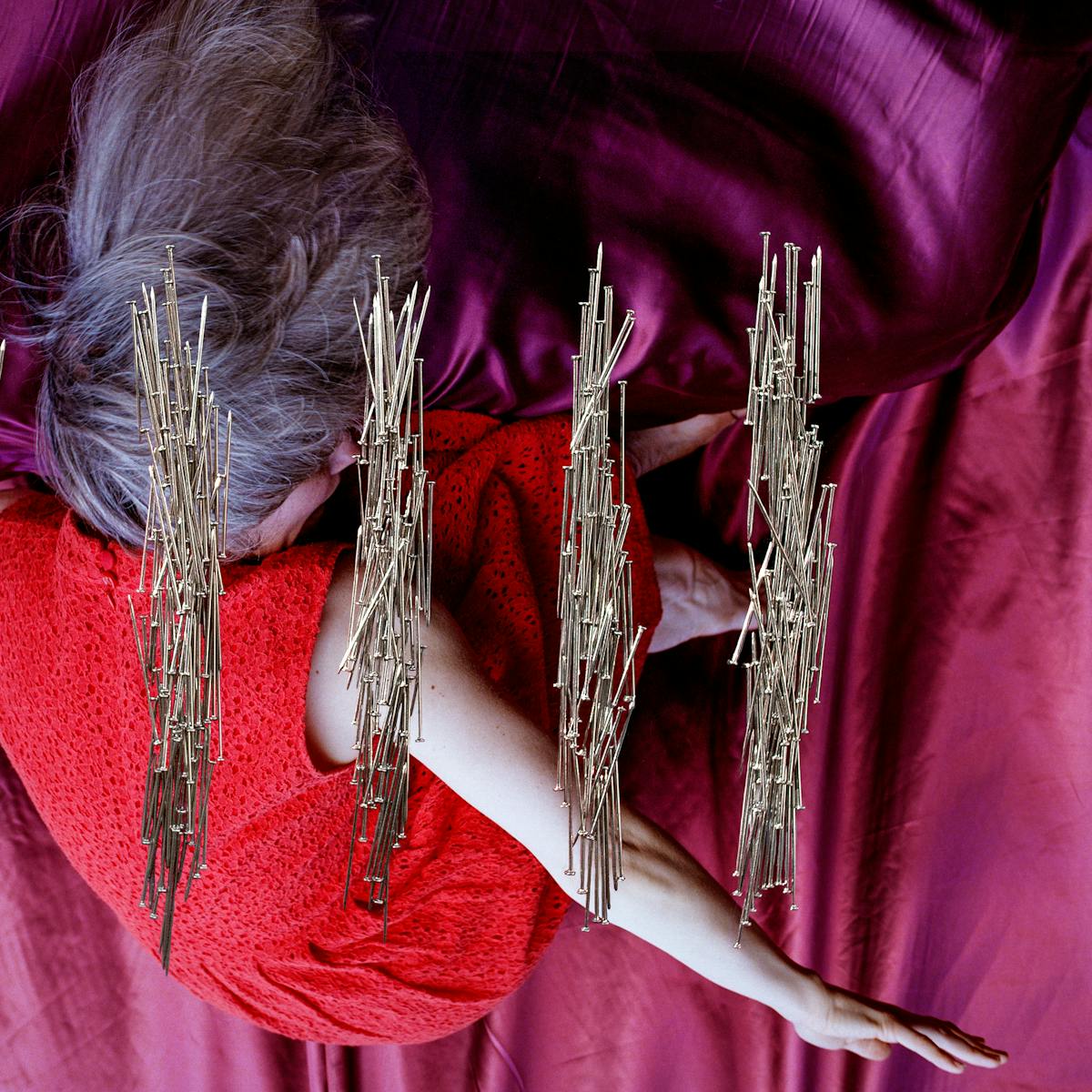 Artwork created with a colour photographic print of a female figure in a bright red dress, set against a purple and blue draped silk background. The figure is on her side, curled up with her knees raised to her chest. Her arms are extended out behind her back, bent at the elbow, with the fingers stretched out. Her body is covered by groups of dress pins, laid on top of the photographic print. The pins are arranged in 5 groups. In each group the pins are arranged vertically to form neat rectangles. They resemble 5 pillars.