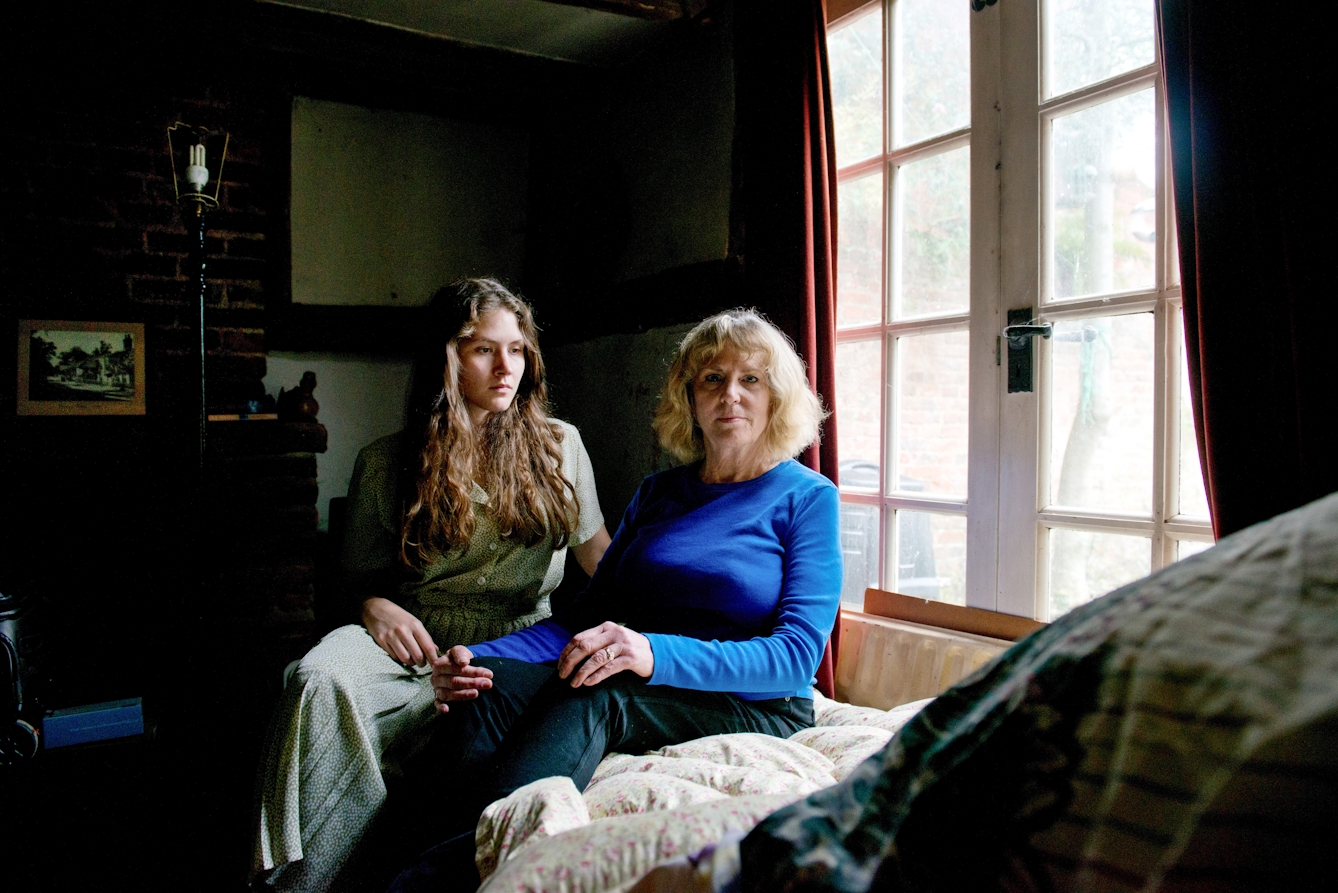 Photograph of a young woman and a mother figure in a domestic room. The young woman is seated in a long dress and and the mother figure sits beside her. The young woman is looking to the mother figure who is looking to camera.