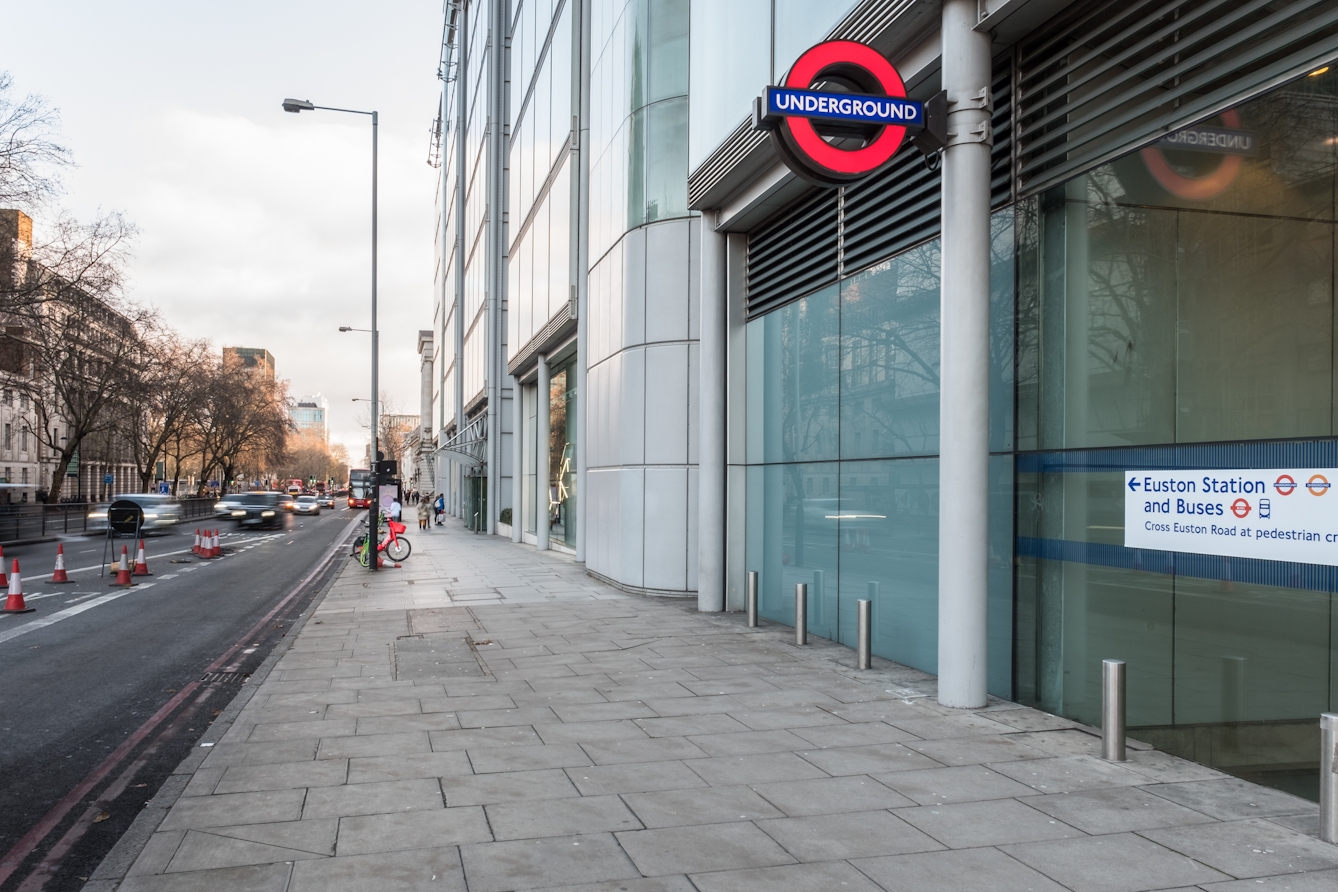 A photograph showing Euston Square underground station on Euston Road at street level with the Wellcome Trust building beyond it, and the Wellcome Collection building beyond that. A pavement runs along all three without the interruption of roads.