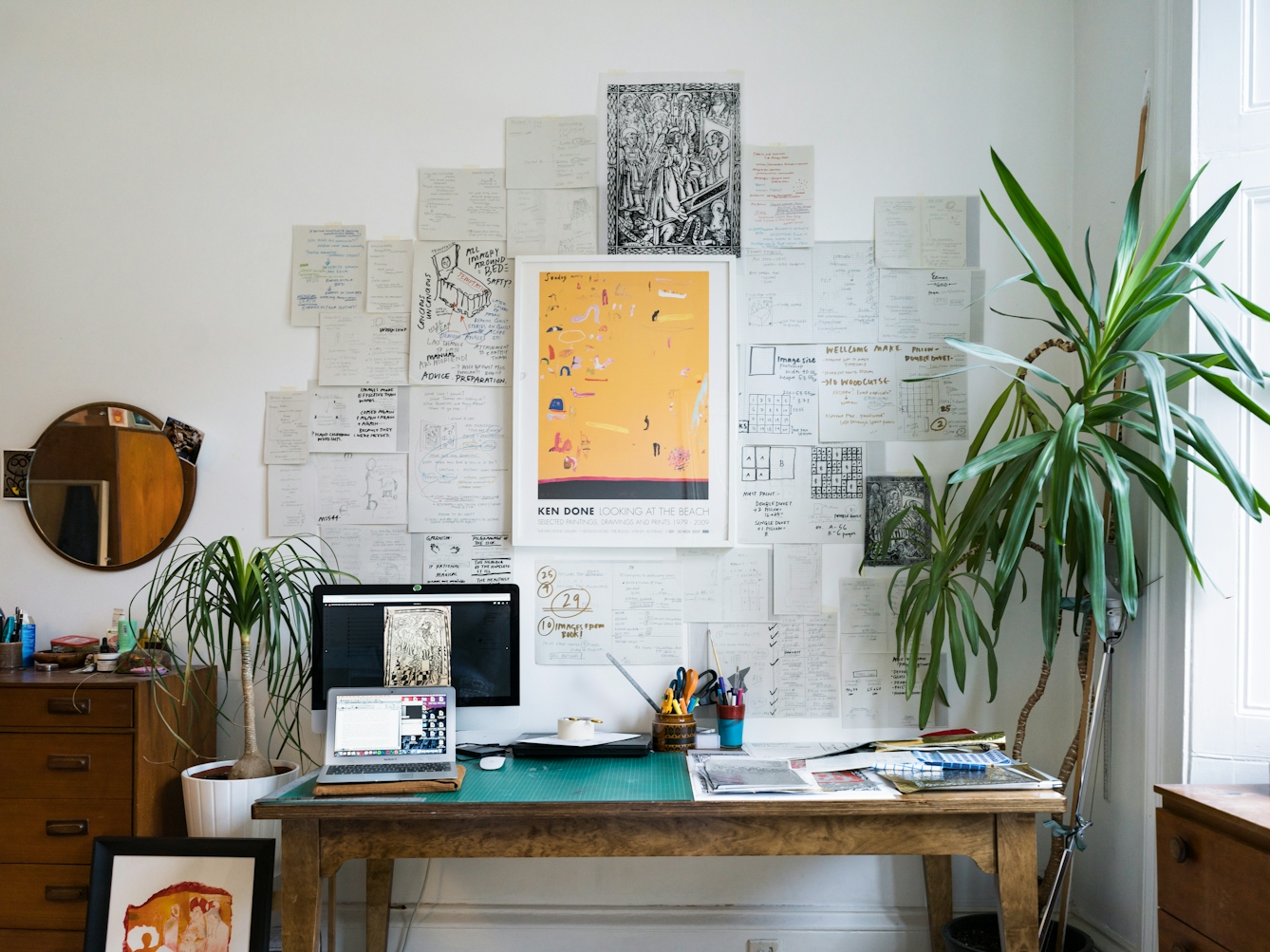 Photograph of a wall covered in pieces of paper which contain sketches and drawings. In front of the wall is a desk on which is a computer screen and laptop, a cutting mat and pen pot holders. Tot he right of the desk is a large pot plant and to the left is a chest of drawers.