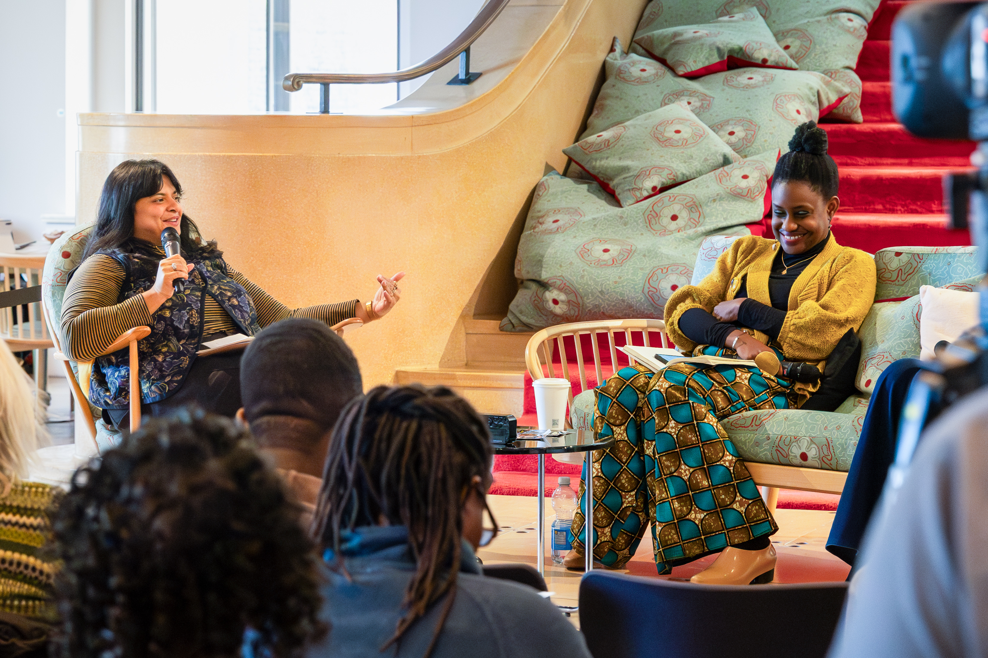 Two people sit together in a relaxed discussion space. The person on the left, holds a microphone and gestures while speaking.