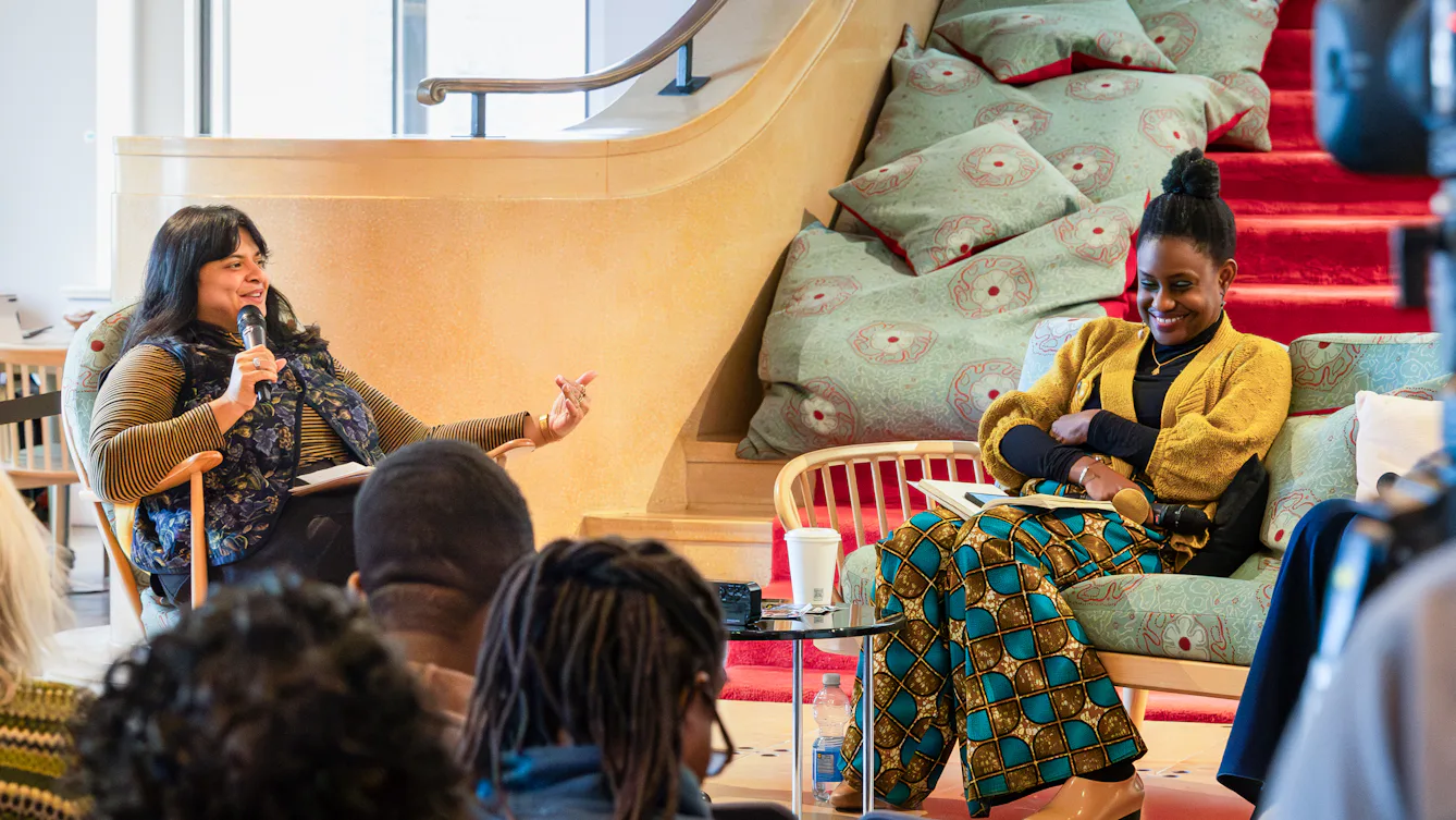 Two people sit together in a relaxed discussion space. The person on the left, holds a microphone and gestures while speaking.