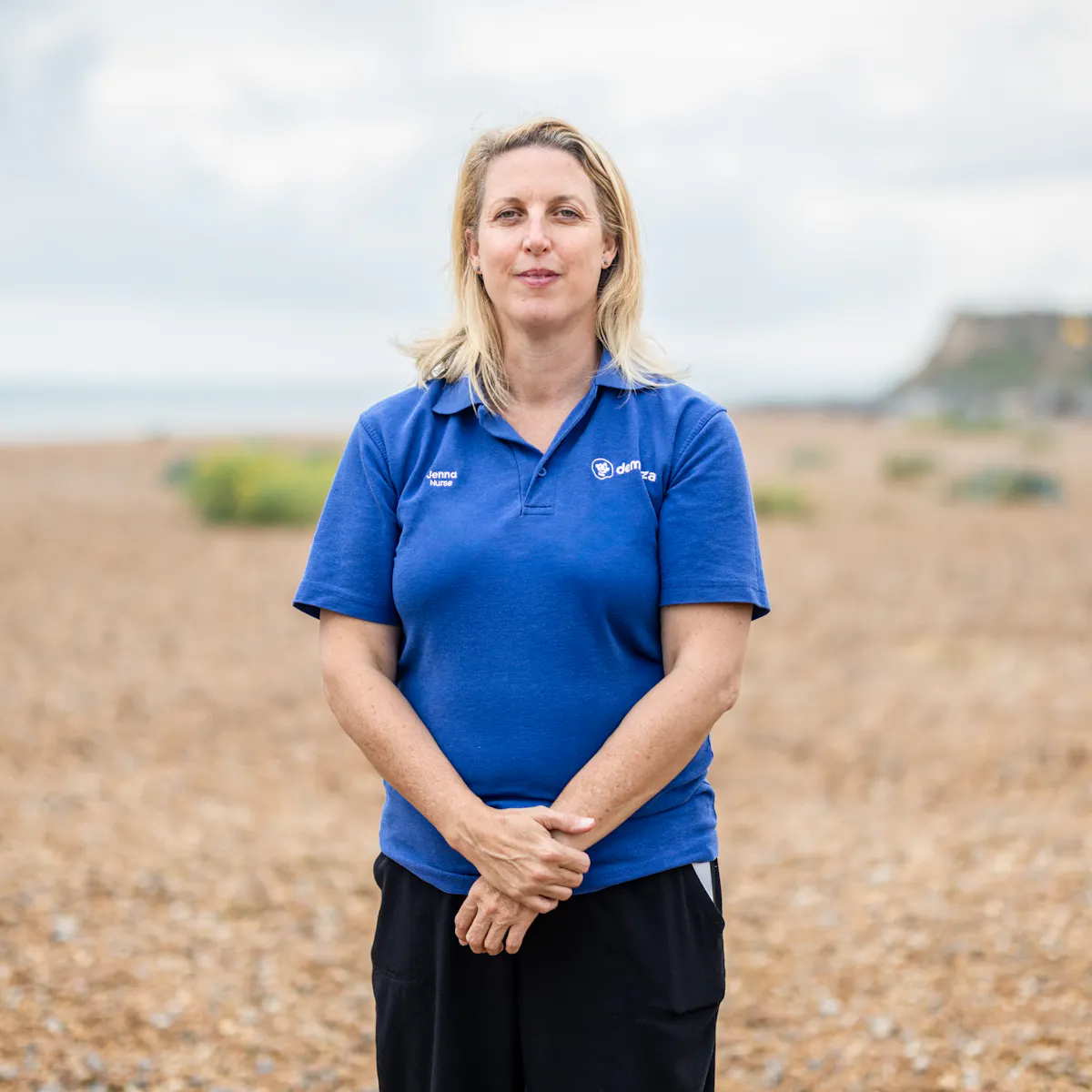 A palliative care nurse in blue polo shirt uniform stands on a pebble beach, hands clasped across her waist, looking straight to camera.