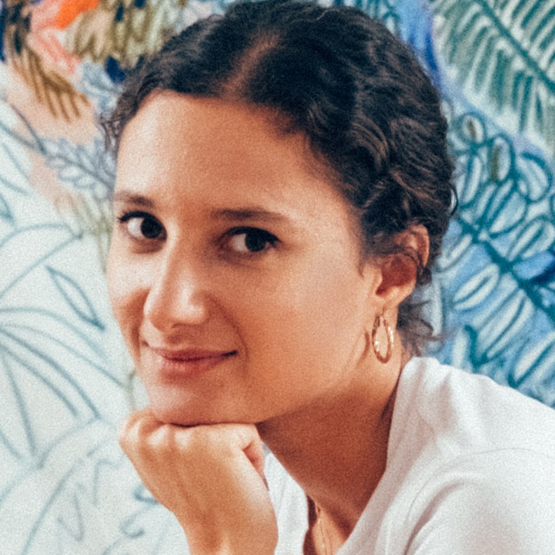 A person with short curly hair rests their chin on their hand and looks towards the camera, smiling softly, with a colourful, abstract, plant-like pattern blurred in the background.