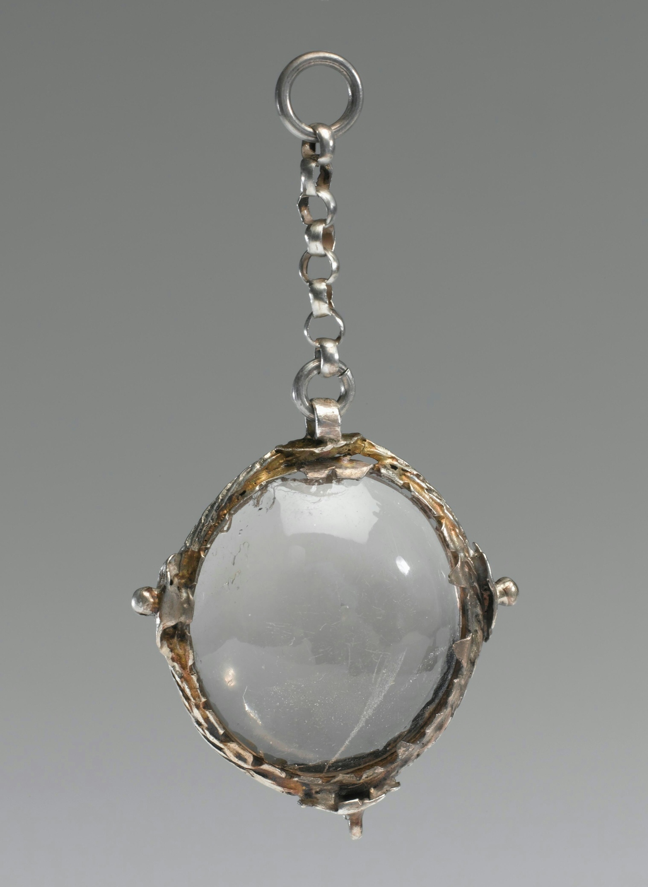 Photograph of a polished oval crystal set in a silver frame, hanging from a short chain, against a grey background.