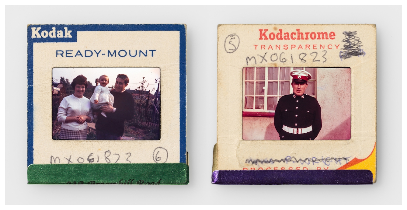 Photograph of two colour 35mm transparencies side by side, each mounted in a cardboard side holder, resting on a white background. The transparency on the left shows a young baby girl in the arms of her father who is stood next to her. They re stood in an urban back garden. The slide mount has a blue border around the edge with Kodak Ready-mount printed on it. A hand written note on the mount reads, "MX061823, 6". The transparency on the right shows the same father this time stood in front of the windows of a house. He's wearing a uniform comprising a black jacket with brass buttons, a thick white belt and a white cap with a red band. The slide mount has the words 'mKodachrome transparency' printed on it. A hand written note on the mount reads, "MX061823, 5".