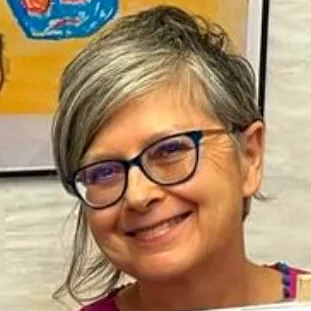 A person with short, wavy, dark hair, wearing a bright top, is smiling to camera and positioned in front of a wall with colourful framed artwork.