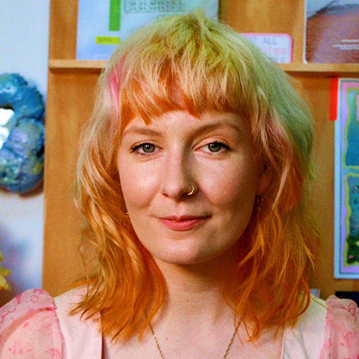 A person with shoulder-length, wavy hair, wearing a light pink, semi-sheer top, positioned in front of a wall with colourful artworks and posters.