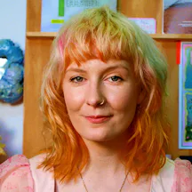 A person with shoulder-length, wavy hair, wearing a light pink, semi-sheer top, positioned in front of a wall with colourful artworks and posters.