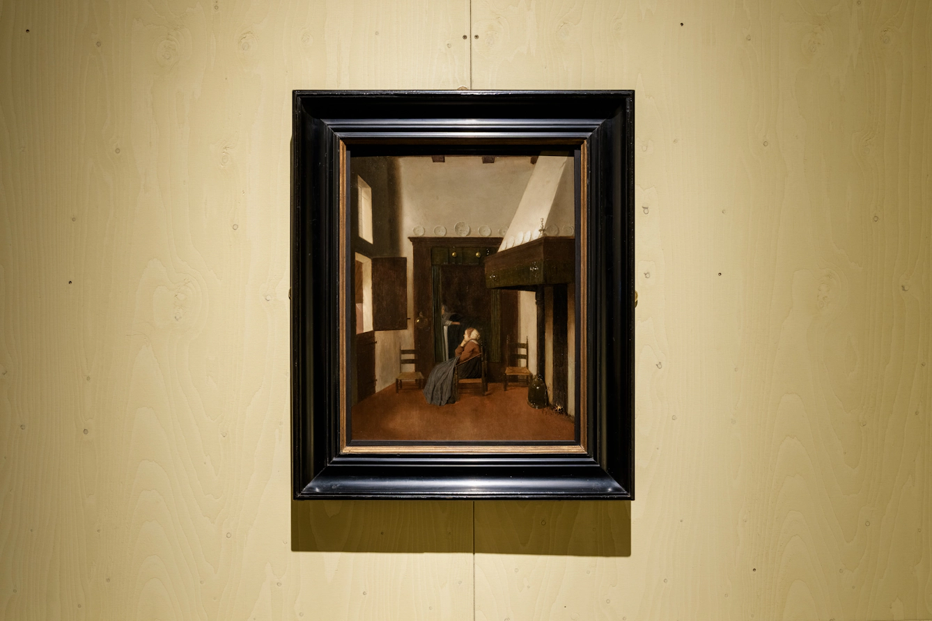 A woman sits by sickbed in a sparse room. There are two empty seats either side of her. 