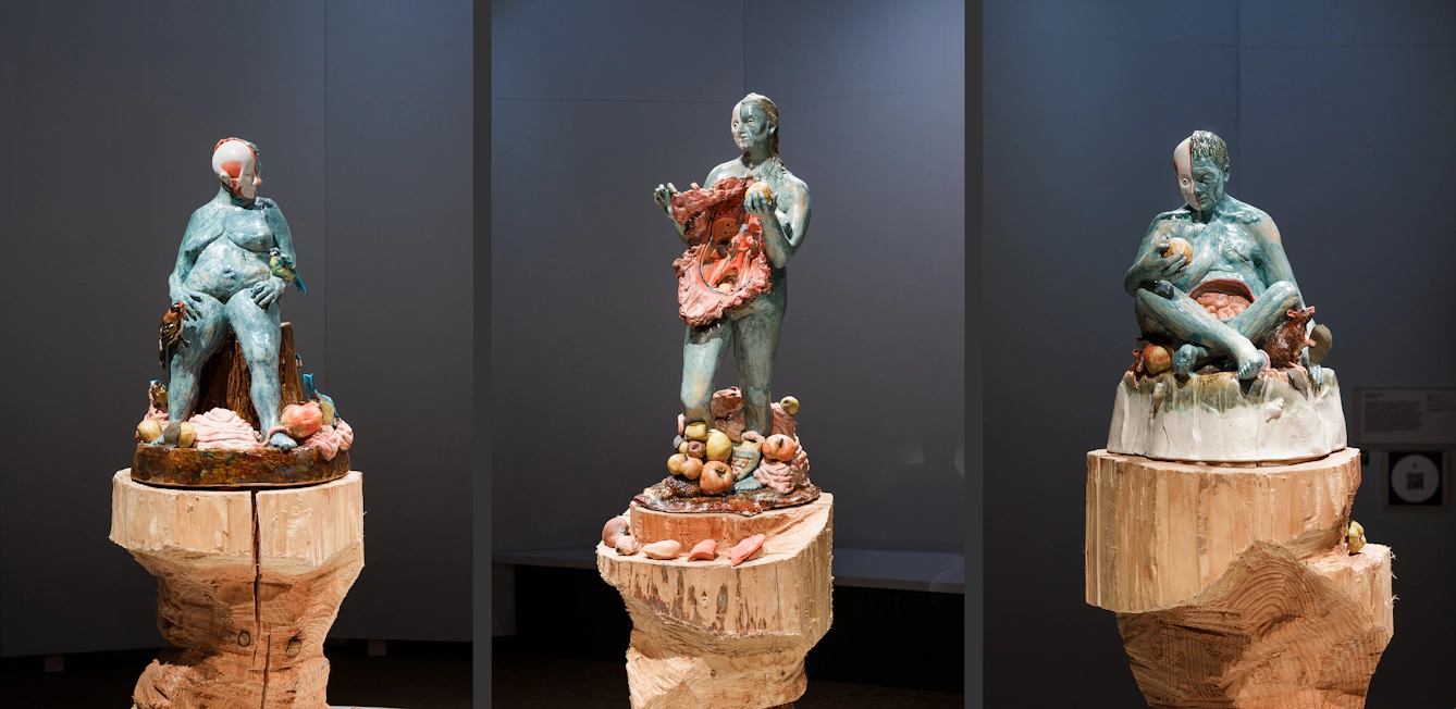 Sculptures of three women on large wooden pillars, surrounded by apples. The central figure's body has been opened up and her insides are visible. 