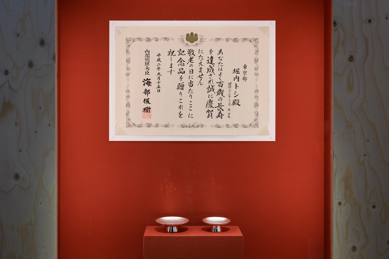 Two metal sake cups on a red stand. Above them is a framed Japanese centenarian certificate congratulating someone on reaching 100 years of age.
