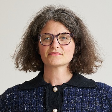 A person with shoulder-length wavy hair, wearing a dark textured jacket with a contrasting collar and two round buttons, faces the camera against a plain light background.