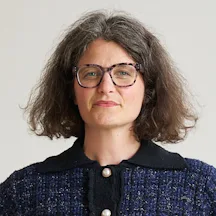 A person with shoulder-length wavy hair, wearing a dark textured jacket with a contrasting collar and two round buttons, faces the camera against a plain light background.