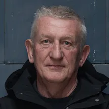A person with short grey hair, wearing a dark jacket, is shown from the shoulders up, standing against a dark, flat background and facing the camera.