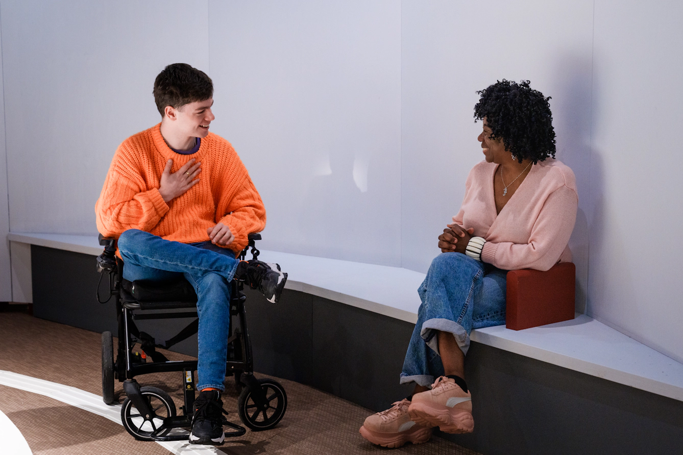 Two visitors talk together. One of them is in a wheelchair, the other is sitting on a bench inside the exhibition.