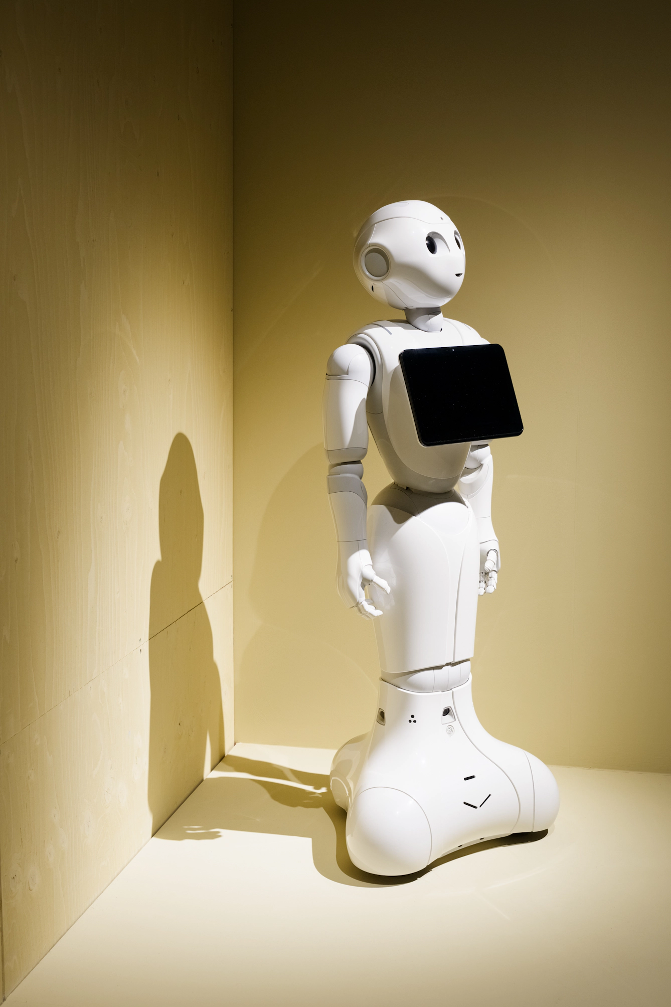 A white robot with a black screen on its chest stands against a yellow wall.
